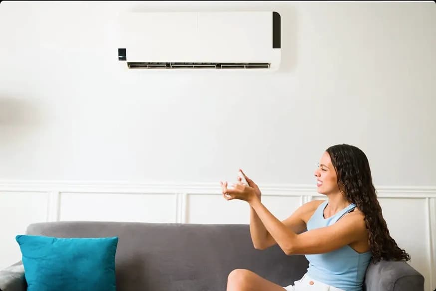Woman feeling warm sitting on sofa looking frustrated adjusting air conditioner that is not cooling properly